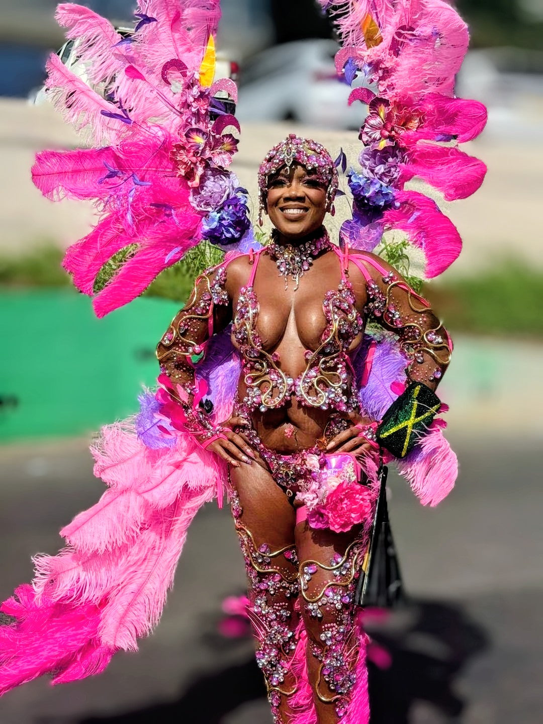 A woman wearing an elaborate carnival costume with bright pink feathers, rhinestone-embellished brown and pink bodysuit, matching feathered headpiece, and a black clutch bag, standing outdoors.