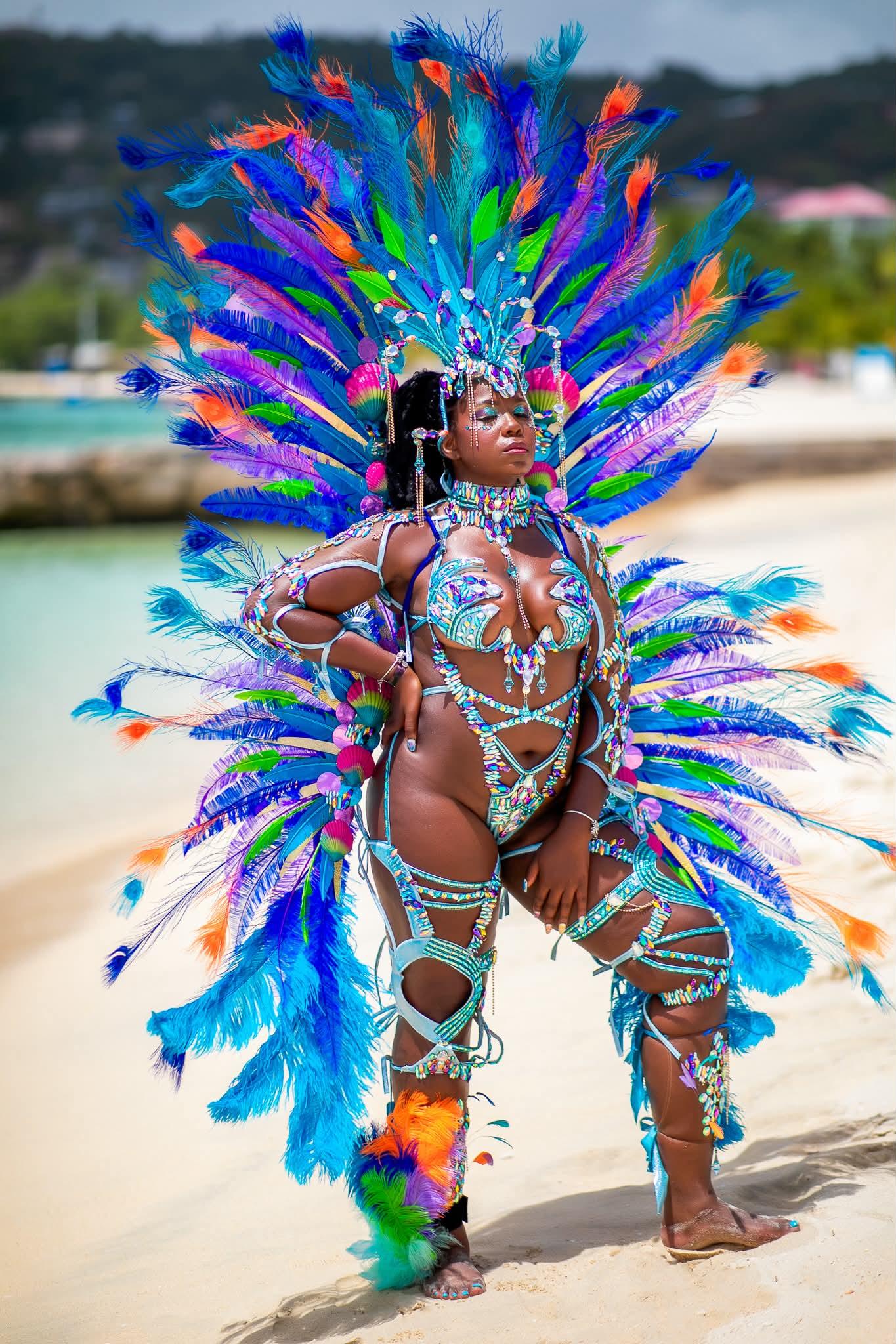 A woman posing on a sandy beach wearing an elaborate carnival costume with a large feathered headdress and matching feathered backpiece, embellished with turquoise, blue, purple, orange, and green feathers and silver beadwork.
